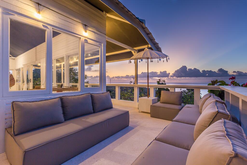 A spacious terrace features comfortable seating arrangements positioned to overlook a scenic view. Soft lighting enhances the area during sunset, while an umbrella provides shade. Large windows enable unobstructed views of the surrounding landscape, creating a serene space for relaxation.