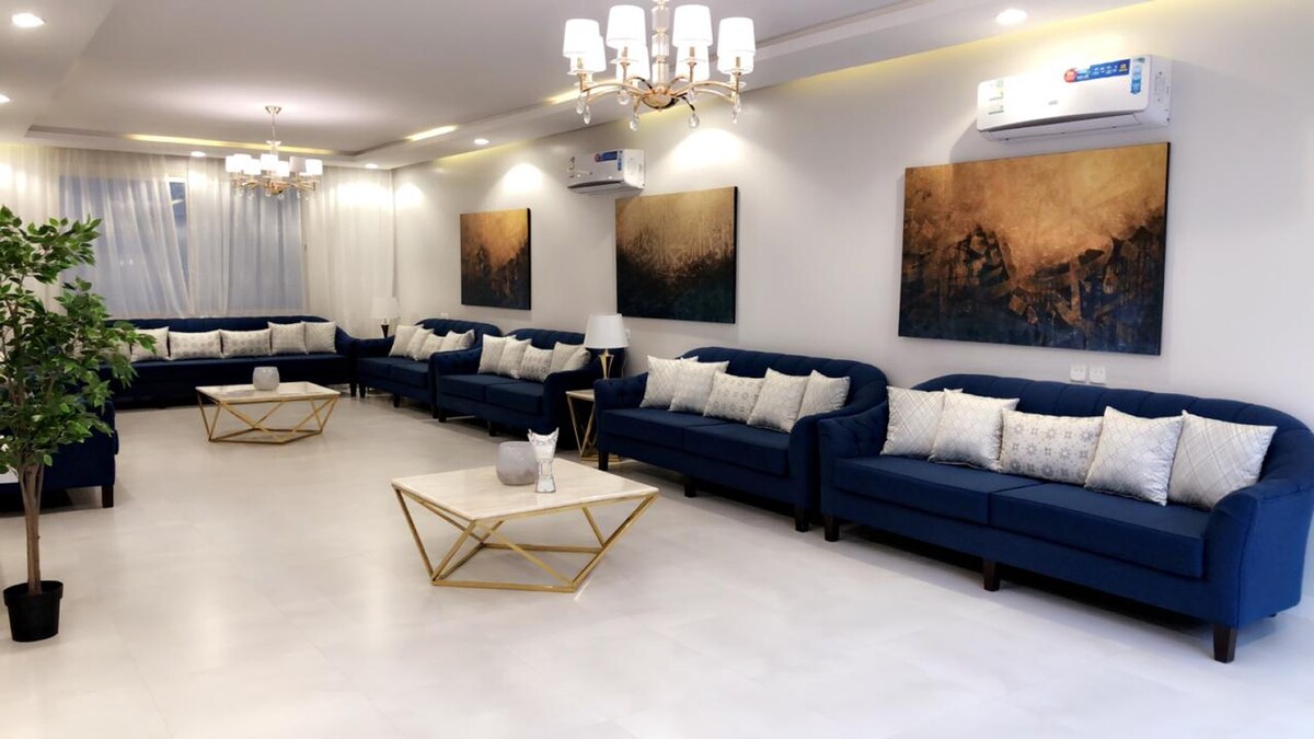 A spacious living area features several navy blue sofas adorned with decorative pillows. Two abstract paintings hang on the light-colored walls, and a coffee table with a minimalist design is positioned centrally. Soft lighting from ceiling fixtures creates a bright and inviting ambiance.