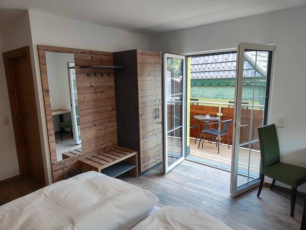 A well-appointed interior features a spacious room with two beds. A wardrobe and bench made from natural wood are seen, along with a large mirror. Bright sunlight enters through glass doors, leading to a private balcony furnished with a small table and two chairs.