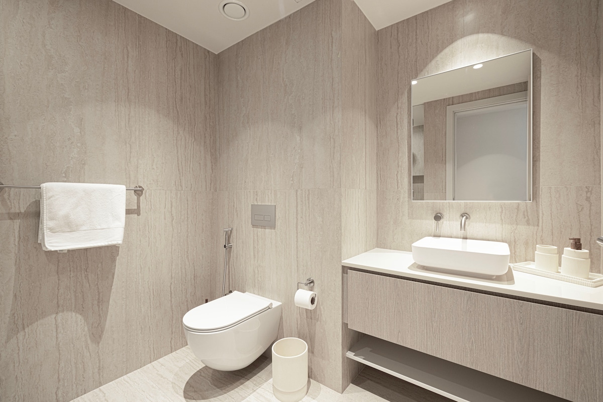 The bathroom features a modern design with light stone walls and flooring. A wall-mounted mirror reflects soft lighting above a sleek vanity with a sink. A toilet and minimalist storage unit are included, along with neatly arranged bath essentials.
