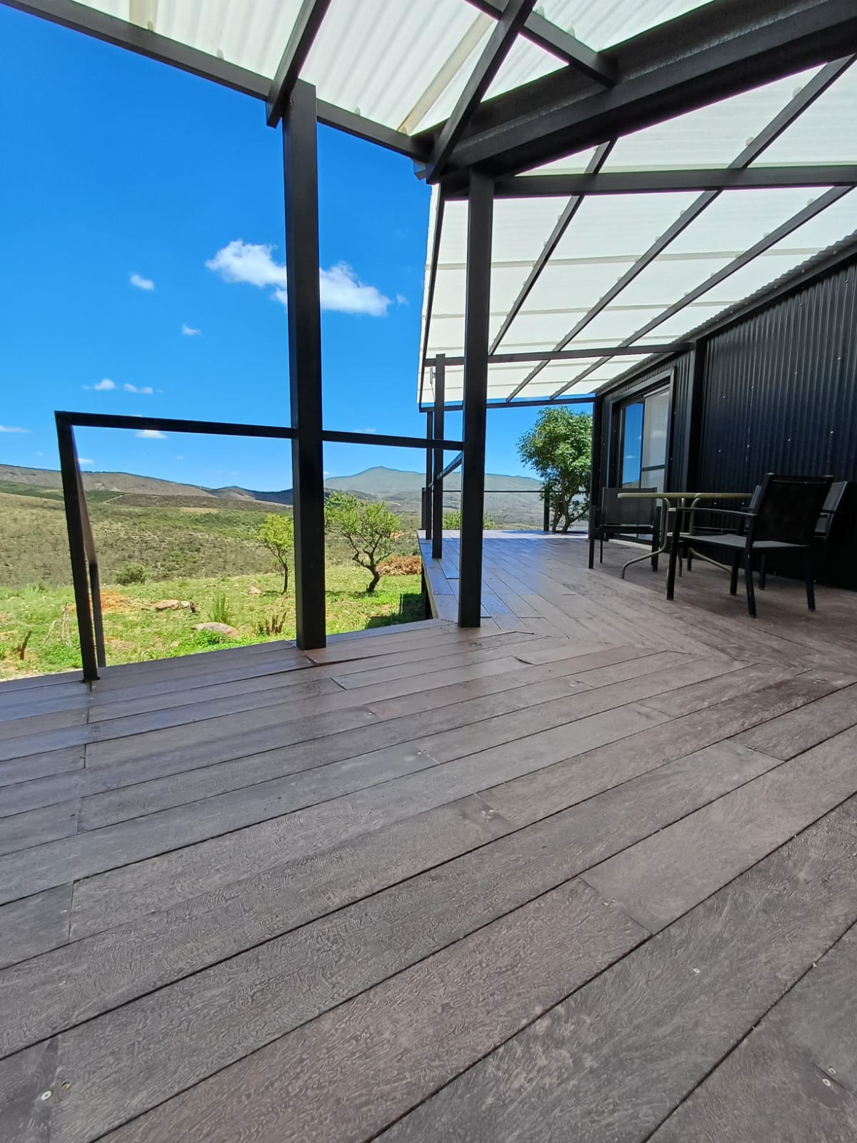 A spacious deck area features wooden flooring and a glass railing, providing an unobstructed view of the surrounding hills and greenery. A clear blue sky is visible, along with a pair of outdoor chairs positioned against the backdrop of nature.
