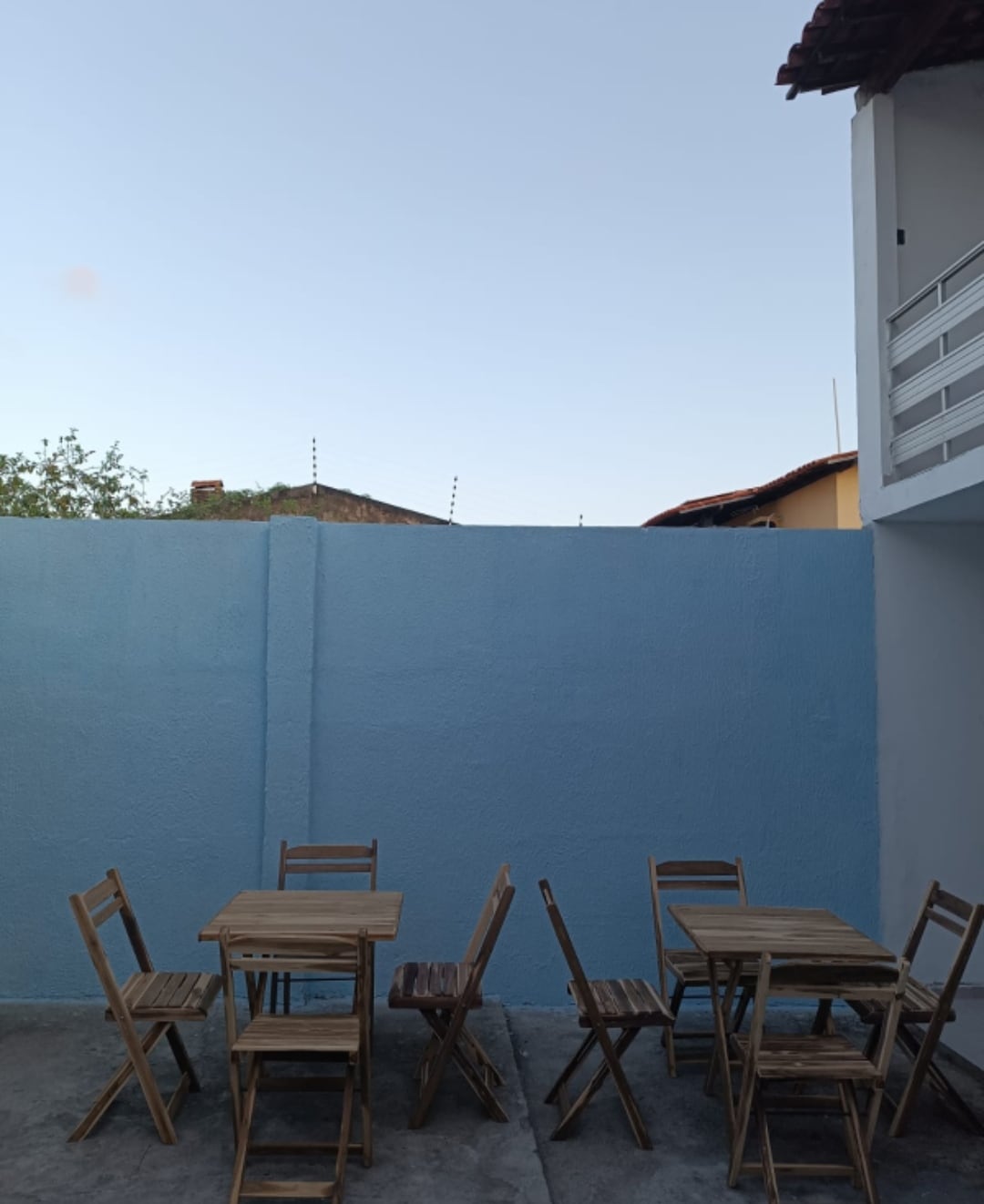 An outdoor area is presented, featuring two wooden tables surrounded by several matching folding chairs. A light blue wall provides a serene backdrop, while the clear sky above enhances the open atmosphere of the space.