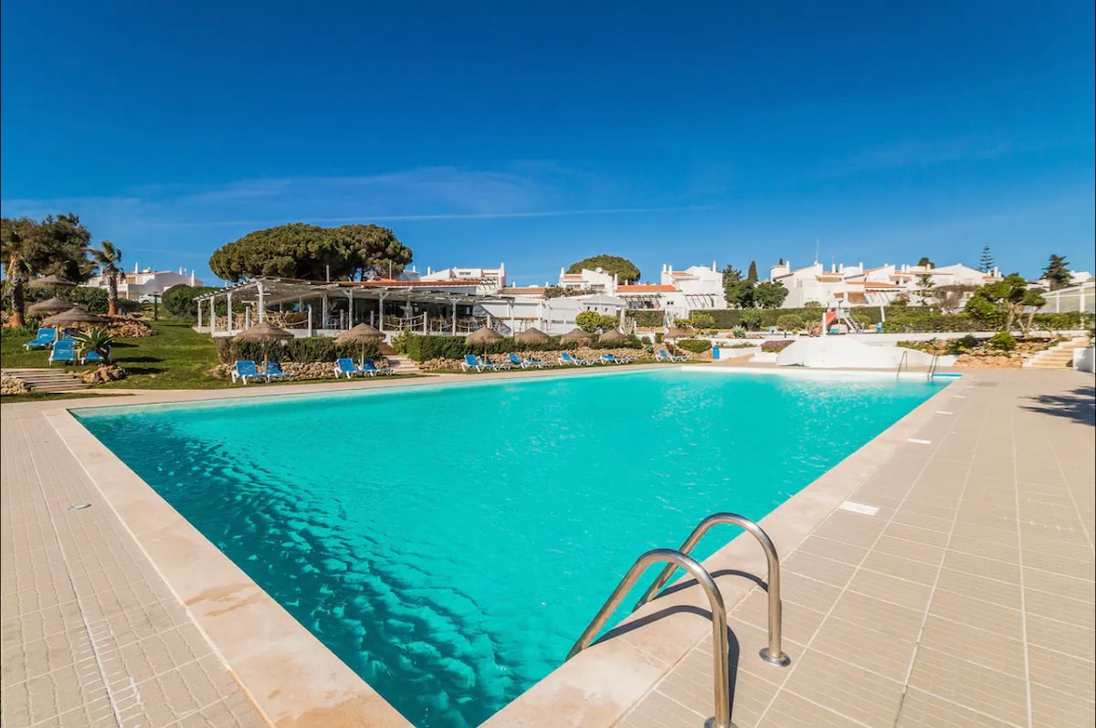 A large rectangular swimming pool is surrounded by textured tiles, with clear turquoise water reflecting the blue sky. Sun loungers line the pool’s edge, and well-maintained greenery is visible in the background, enhancing the serene environment.