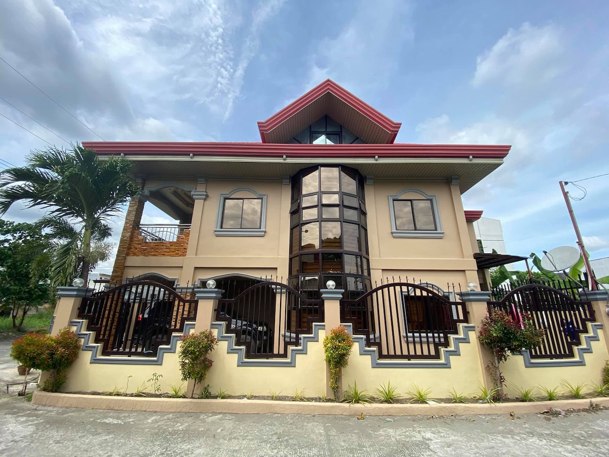 The exterior of the house showcases a two-story design with large glass windows that allow ample natural light. The roof is sharply peaked and has red accents. A gated entrance is flanked by decorative plants, and a spacious terrace is visible on the upper level.