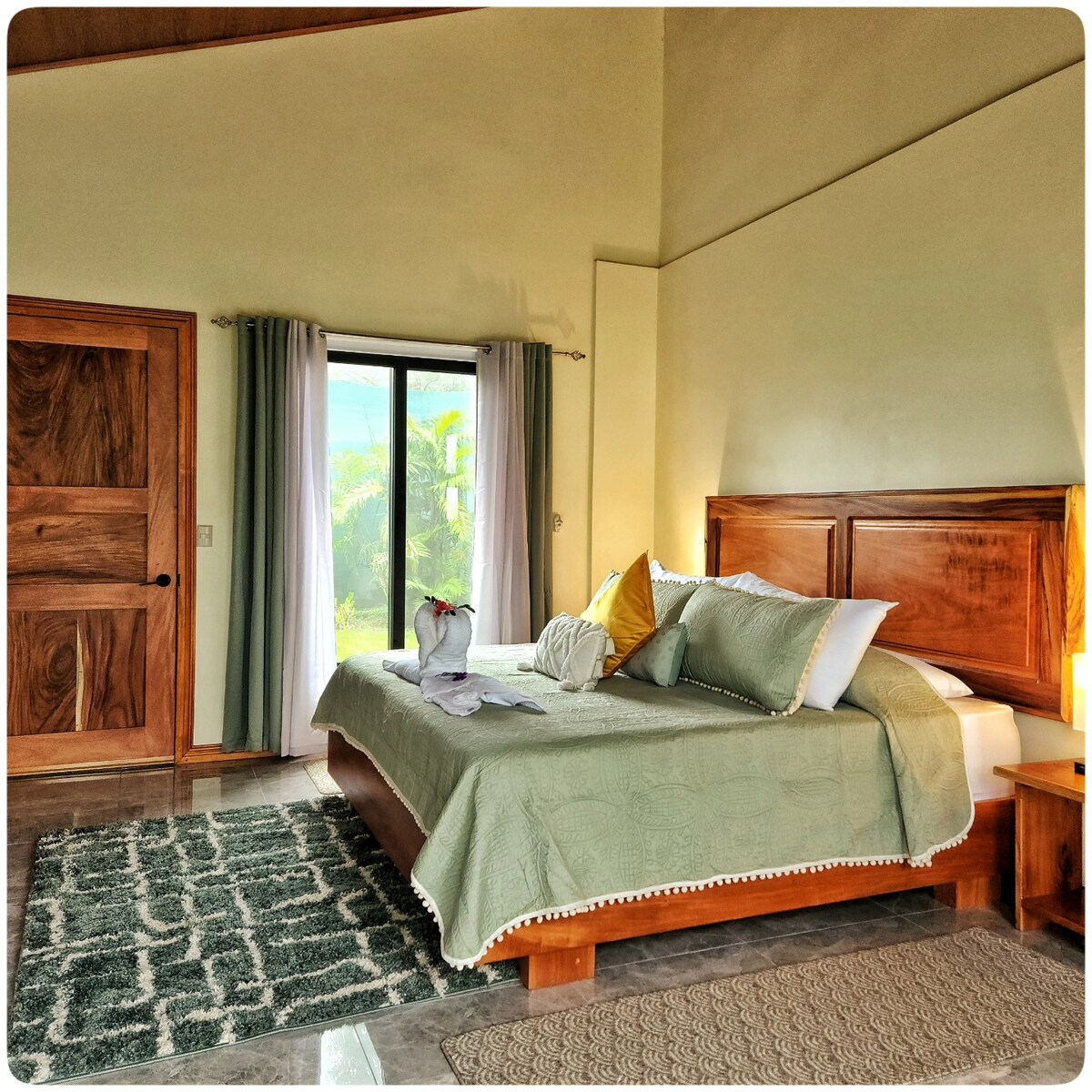 A spacious sleeping area is highlighted by a large bed dressed in a soft green coverlet. Natural light fills the room through a sliding glass door, which offers views of the surrounding greenery. A plush area rug adds texture to the polished floor.