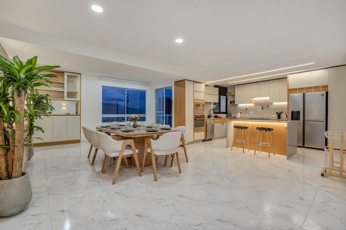 An open kitchen and dining area featuring modern cabinetry and a large dining table with stylish chairs. A kitchen island with bar stools provides additional seating. Natural light enters through large windows, illuminating the polished marble flooring and the contemporary design.