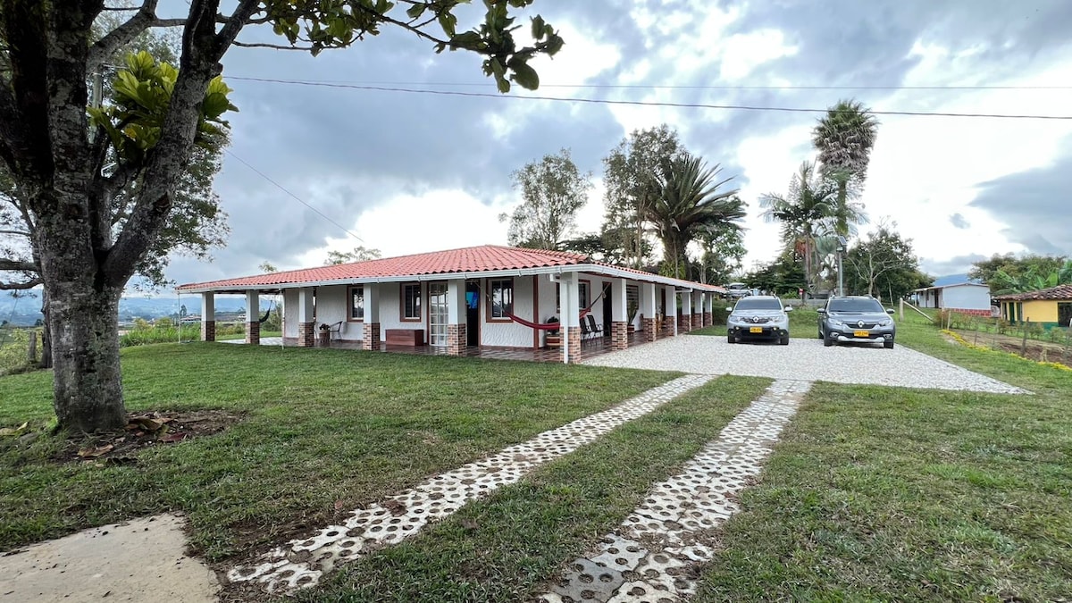 A spacious property is surrounded by lush greenery, featuring a well-maintained lawn and a gravel pathway leading to a charming single-story building. The structure has a red-tiled roof, multiple entries, and parking spaces for two vehicles, creating an inviting outdoor setting.