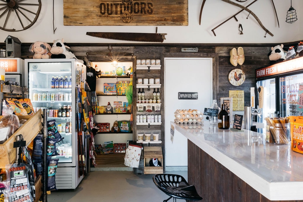 A farm store interior is visible, featuring a wooden counter with assorted goods displayed on top. Shelves hold various snacks and drinks, including a refrigerator filled with beverages. Rustic decor elements enhance the ambiance, while toys and playful items are arranged throughout.