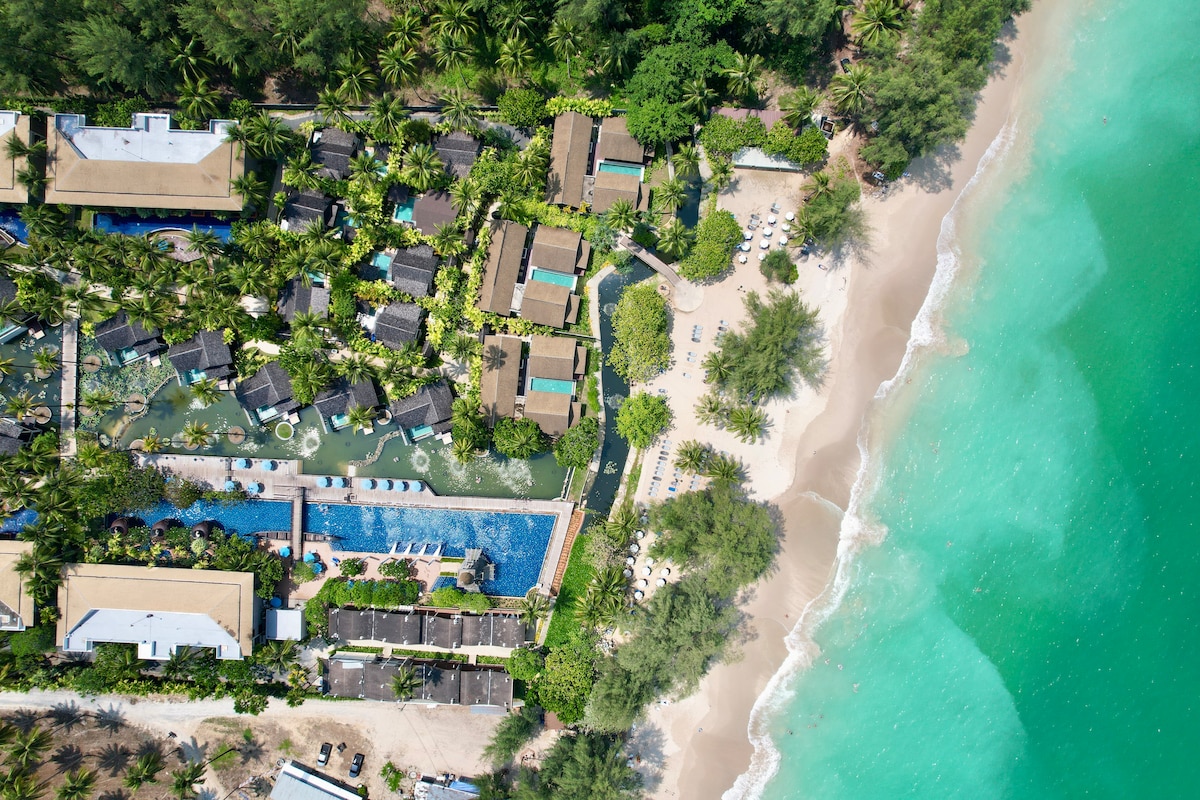 An aerial view captures the resort's layout, showcasing lush palm trees surrounding various accommodations and a large swimming pool. The image highlights proximity to a white sandy beach, with the serene turquoise waters of the Andaman Sea gently lapping at the shore.