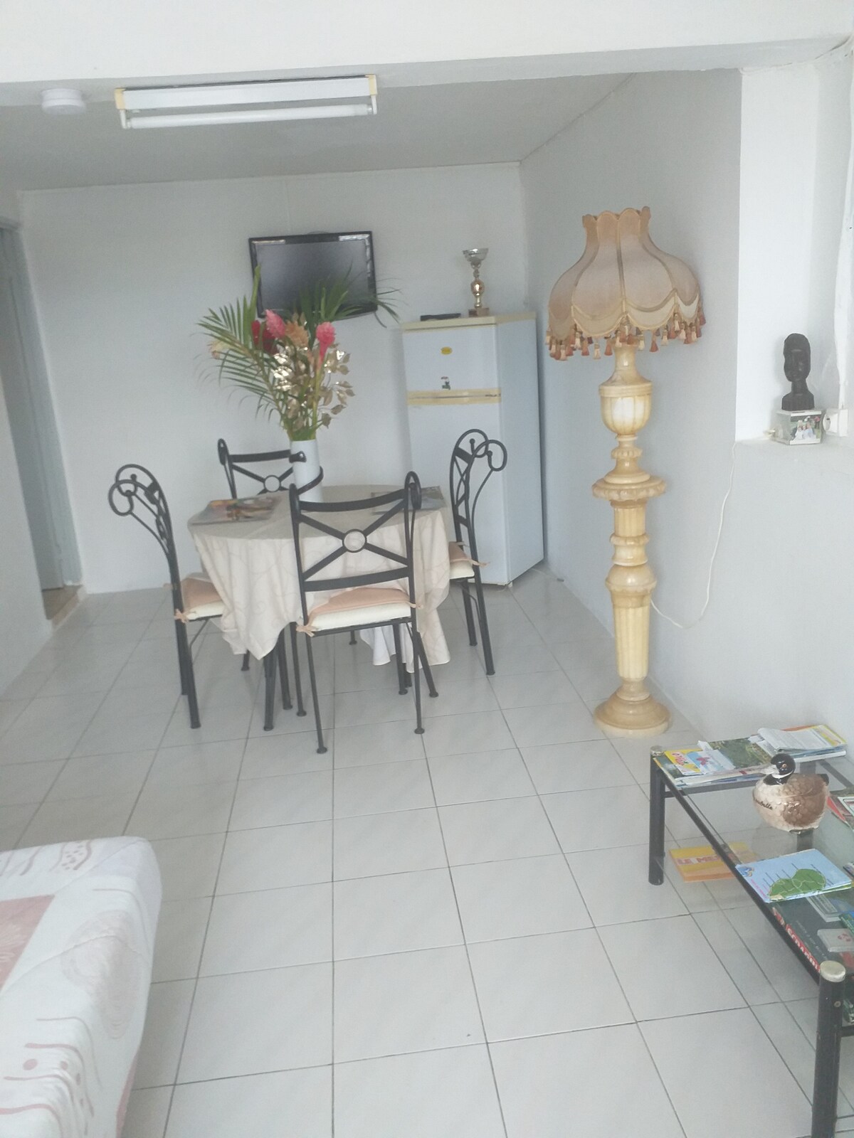 A dining area is presented with a round table surrounded by four black chairs, complemented by a beige tablecloth. A decorative lamp stands nearby, illuminating the space. A small fridge and a television are visible, with a few decorative items placed strategically around the room.