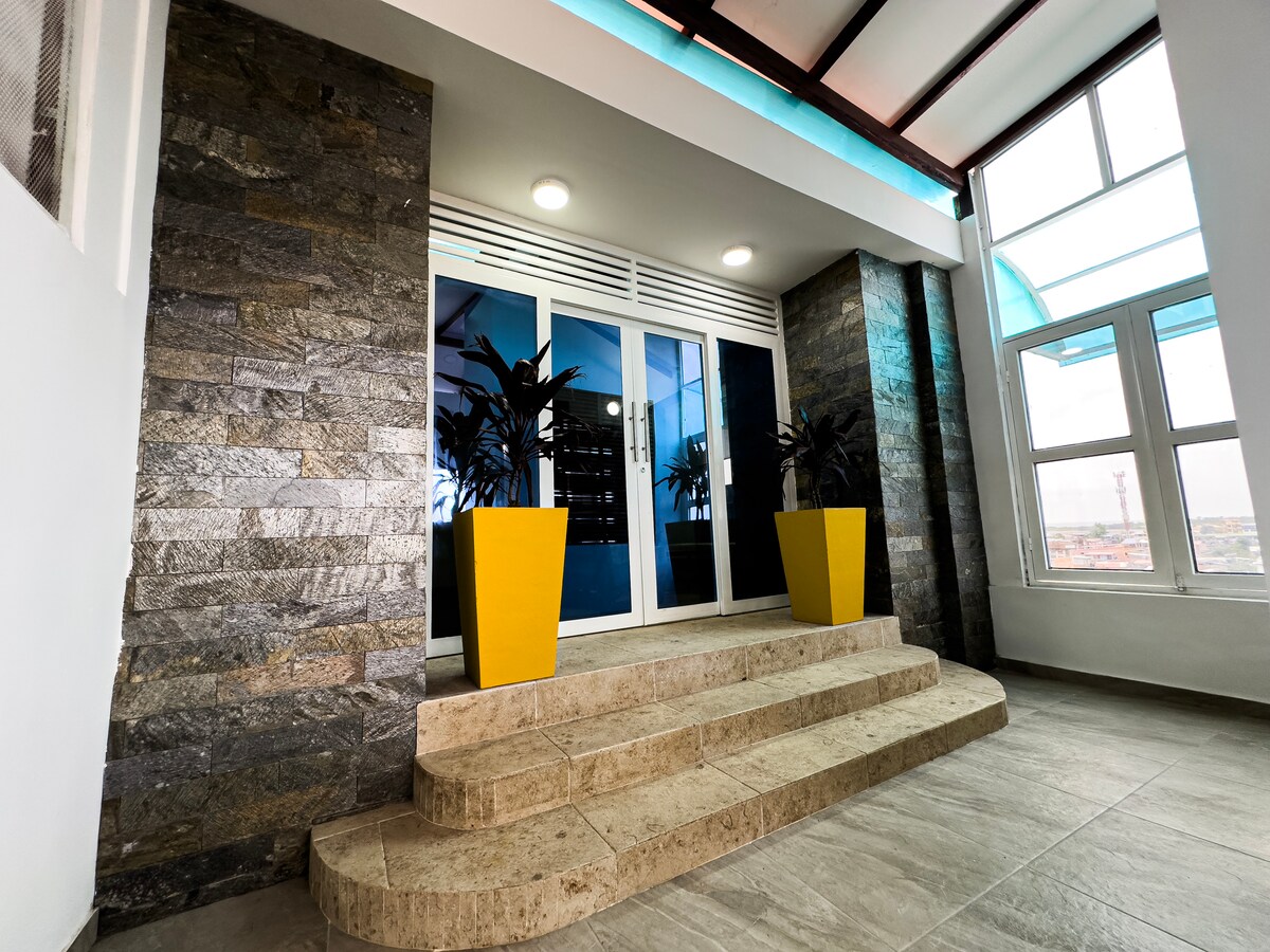 A welcoming entrance is framed by two large yellow planters, filled with lush green plants. Stairs lead to a glass door, surrounded by textured stone walls and bright overhead lighting, creating a vibrant first impression.