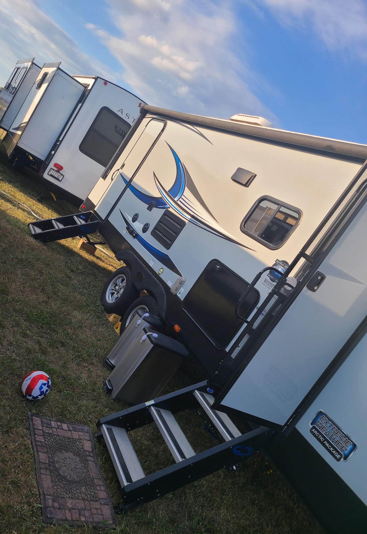 A clean and modern camper is positioned on a grassy area, displaying a sleek exterior design with blue accents. Steps lead up to the entry door, and a small area rug is placed near the base of the stairs. A football rests on the ground nearby.
