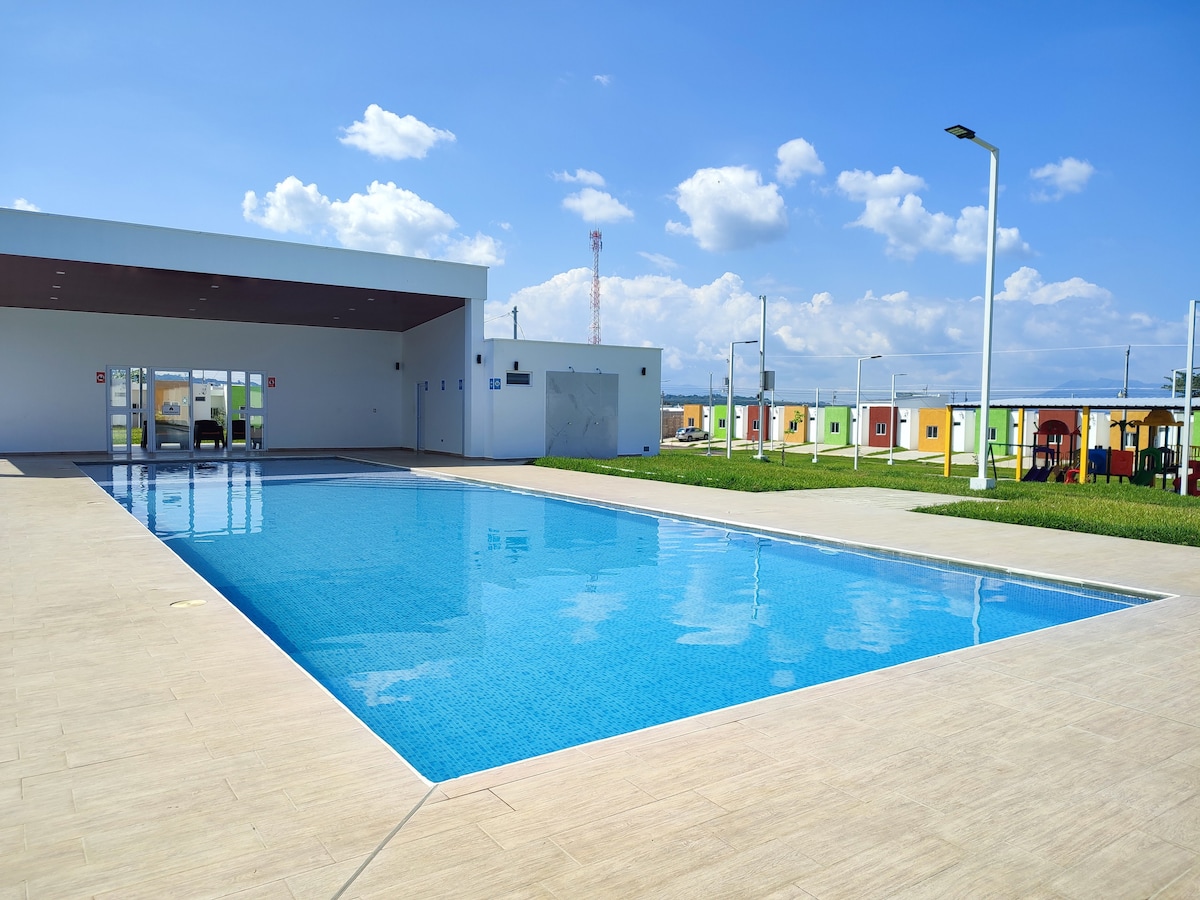 A clear blue swimming pool sits in a well-maintained area, reflecting the sky above. Adjacent to the pool, a shaded area features spacious seating. Colorful structures are visible in the background, along with a children’s play area. Green grass frames the elegant poolside.