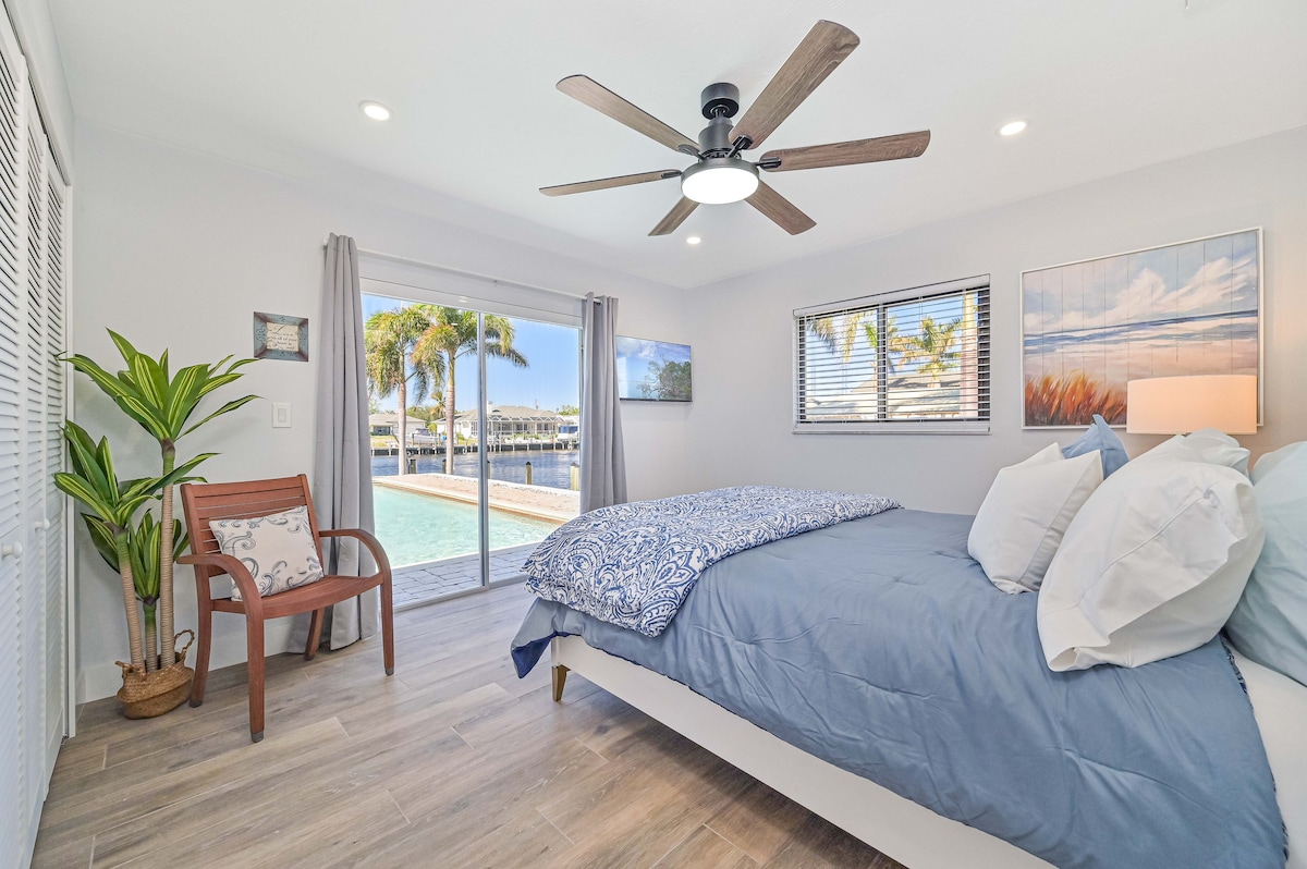 A comfortable bedroom features a spacious bed with blue bedding, accompanied by a decorative plant and a wooden chair. Natural light floods the room through sliding glass doors that open to a pool and waterfront view.
