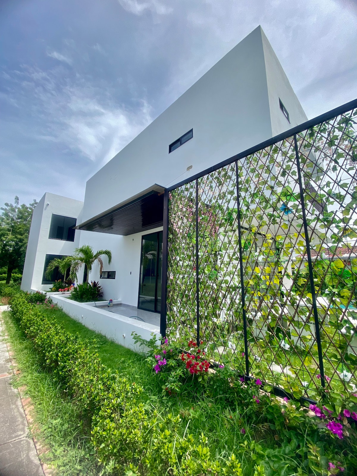 The exterior of the villa showcases a modern white façade, complemented by a lush green garden with flowering plants. A decorative lattice with climbing vines adds a touch of elegance, creating a welcoming entrance next to the path.