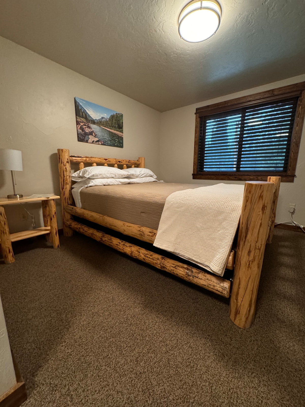 A rustic bedroom features a log-frame bed with a neutral-colored blanket. A bedside table with a lamp is positioned to the left. Natural light enters through a window dressed with wooden blinds, and a mountain landscape photograph hangs on the wall.