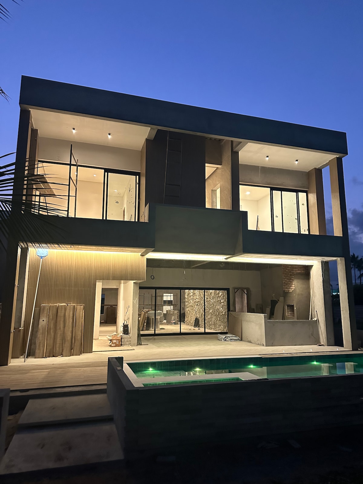 The modern exterior of the newly constructed home showcases clean lines and large glass doors that invite natural light. A private pool with subtle illumination is visible in the foreground, enhancing the inviting outdoor space. The structure is enveloped by a serene evening sky.