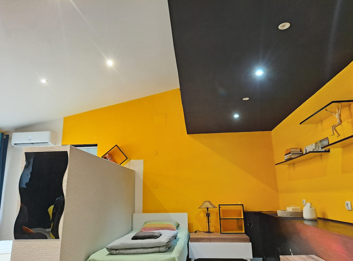A bright room features a sloped ceiling with recessed lighting. One side is painted in a vibrant yellow hue, contrasting with the dark ceiling. A spacious bed is positioned in the corner, accompanied by a bedside lamp and a minimalist shelf displaying decorative items.