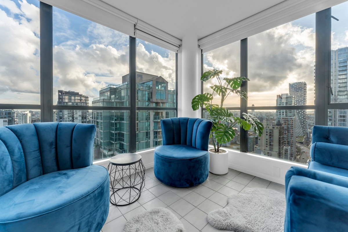 A corner seating area features two plush, blue chairs facing large windows that provide expansive views of the city skyline. A small, round table is positioned alongside the chairs. A potted plant adds a touch of greenery to the modern space, illuminated by natural light.