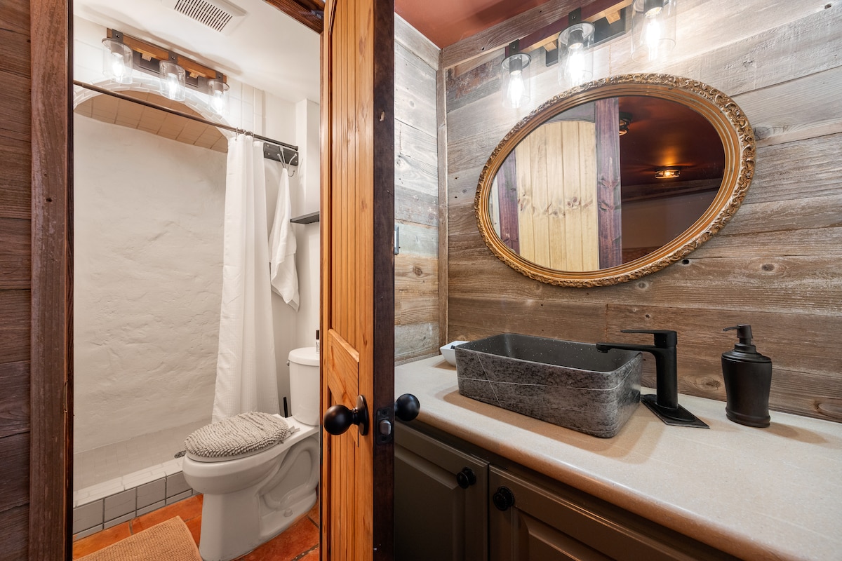 A bathroom is featured with rustic wooden accents and soft lighting. A dark stone sink is paired with a modern faucet. The space includes a toilet and a shower area, which is enclosed by a curtain. The warm tones enhance the overall ambiance.