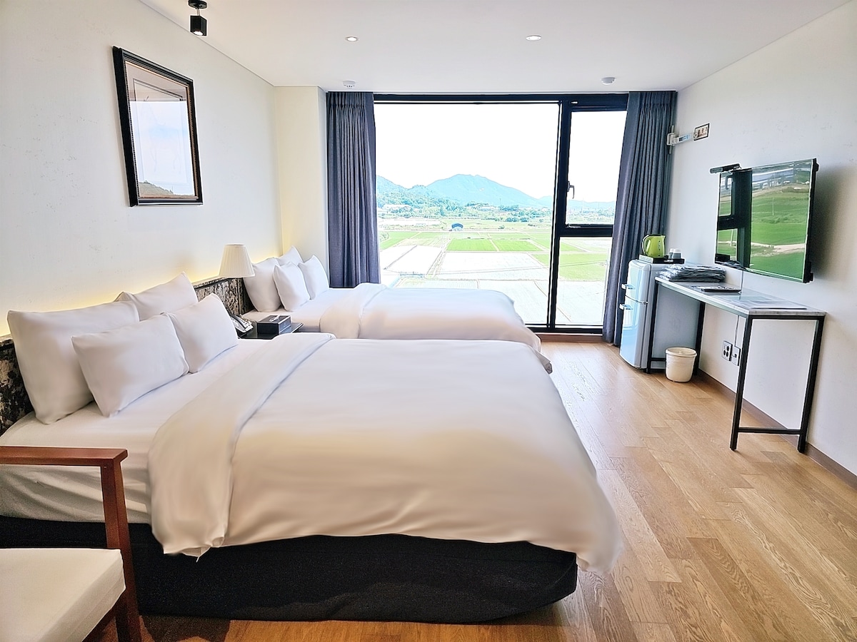A modern guestroom is presented with two spacious double beds dressed in crisp white linens. A large window reveals an expansive view of green fields and mountains. A flat-screen television is mounted on the wall, and a small refrigerator is positioned in the corner.