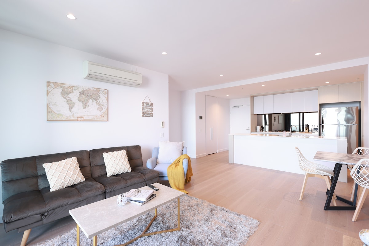 A modern living area is shown, featuring a comfortable sofa with patterned cushions and a decorative throw. A coffee table occupies the center, while a dining area with stylish chairs is visible in the background. Light streams in through large windows, enhancing a bright and airy atmosphere.