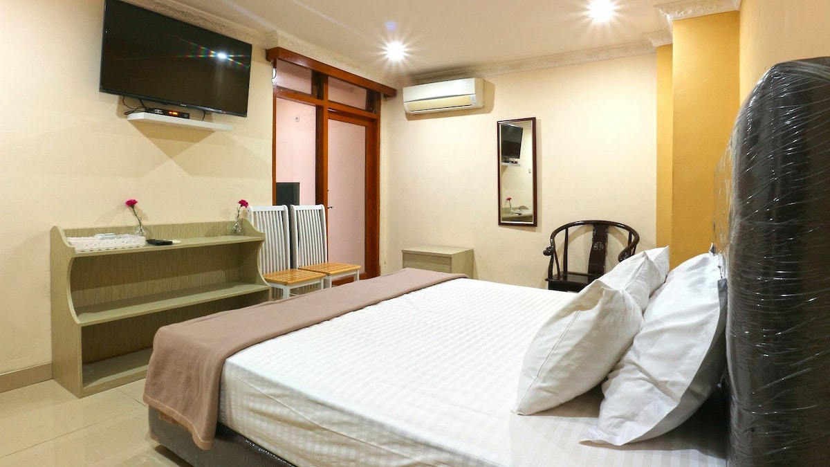 A standard double room features a comfortable bed with neatly arranged white linens. A television is mounted on the wall opposite, with a decorative mirror nearby. A desk and two chairs are positioned against one wall, while air conditioning is visible above.