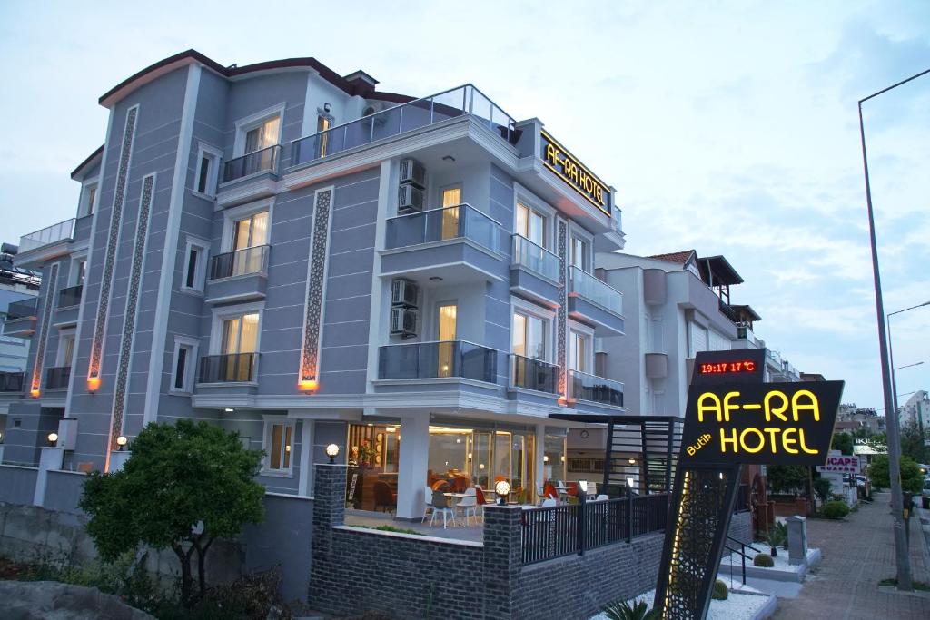 The exterior of AF-RA Hotel is presented, showcasing a modern three-story building with a light grey facade and balconies on the upper levels. The hotel sign is prominently displayed, and an outdoor seating area is visible on the ground floor, surrounded by greenery.