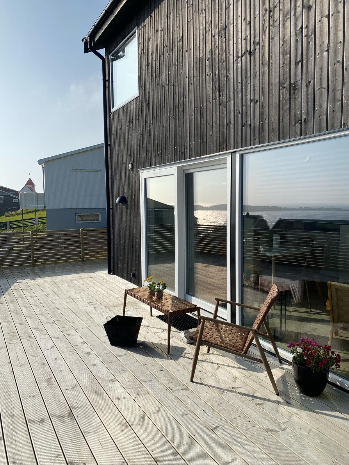 A wooden deck features a small table and two chairs, positioned to enjoy the view of the water. Potted plants add a touch of greenery, while large glass doors provide access to the interior. The surrounding area is peaceful, enhancing the outdoor experience.