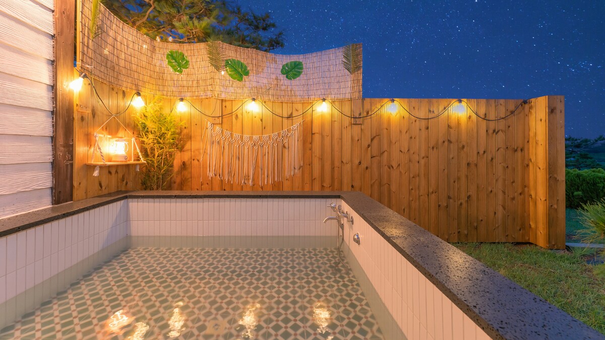 An inviting outdoor hot tub is surrounded by wooden fencing, adorned with fairy lights that create a gentle glow. The mosaic-tiled interior of the tub reflects the starry sky above, enhancing the tranquil ambiance of the secluded space.