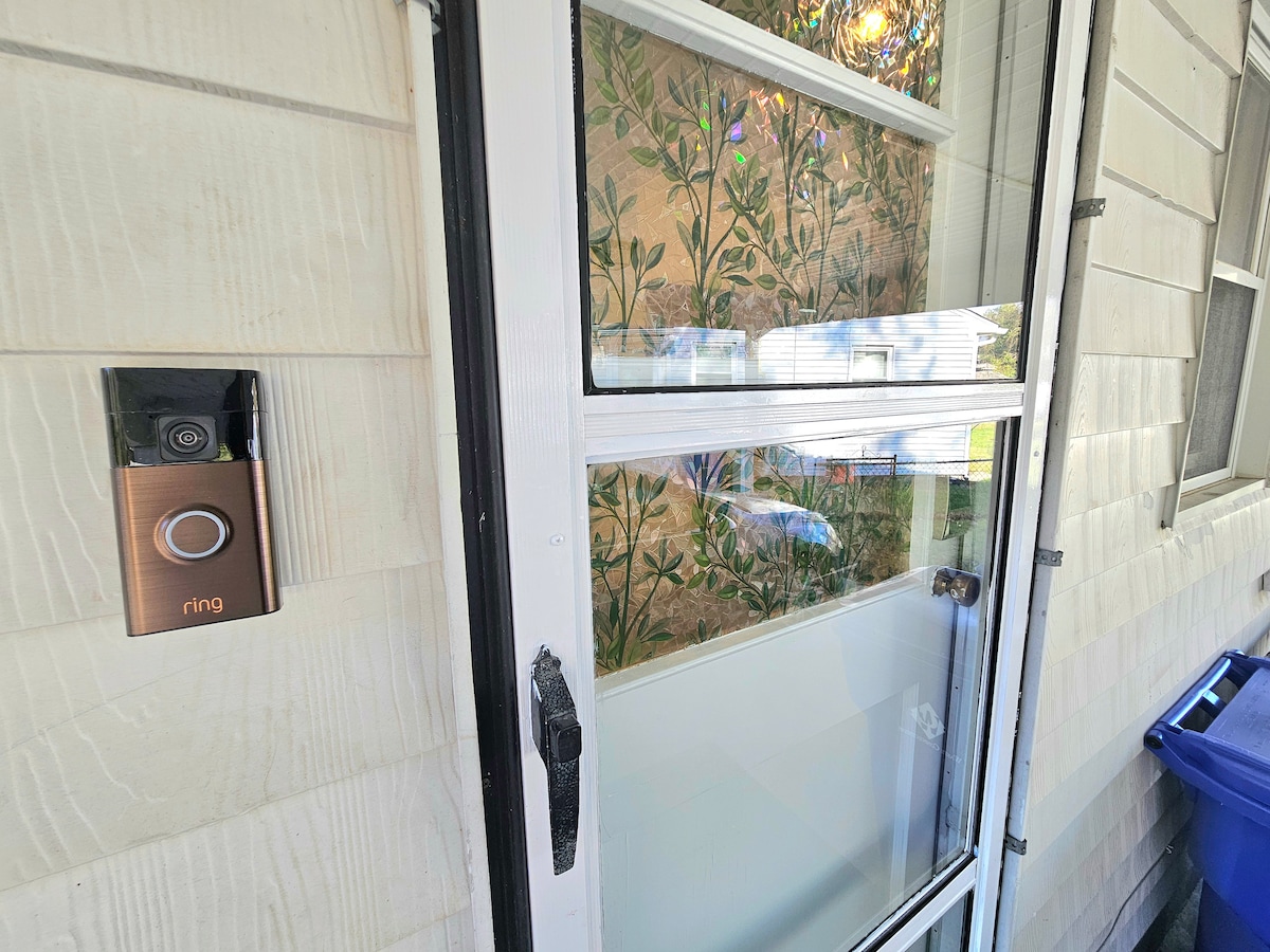 An entryway is presented, featuring a modern doorbell on the side wall alongside a door with a window pane. The door's glass showcases a floral-patterned privacy shade, allowing light to filter through while maintaining a cozy ambiance.