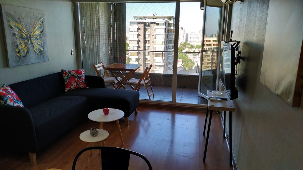 A modern living space features a large window that provides city views, complemented by natural light. A sleek black sofa adorned with colorful cushions is positioned beside a small coffee table. A dining area with a wooden table and chairs is visible, creating a cozy setting.