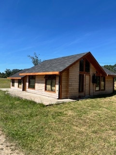 A spacious wooden house is depicted, featuring large windows that allow natural light to fill the interior. The sloped roof adds a charming architectural element, while the surrounding grassy area provides a serene outdoor setting.
