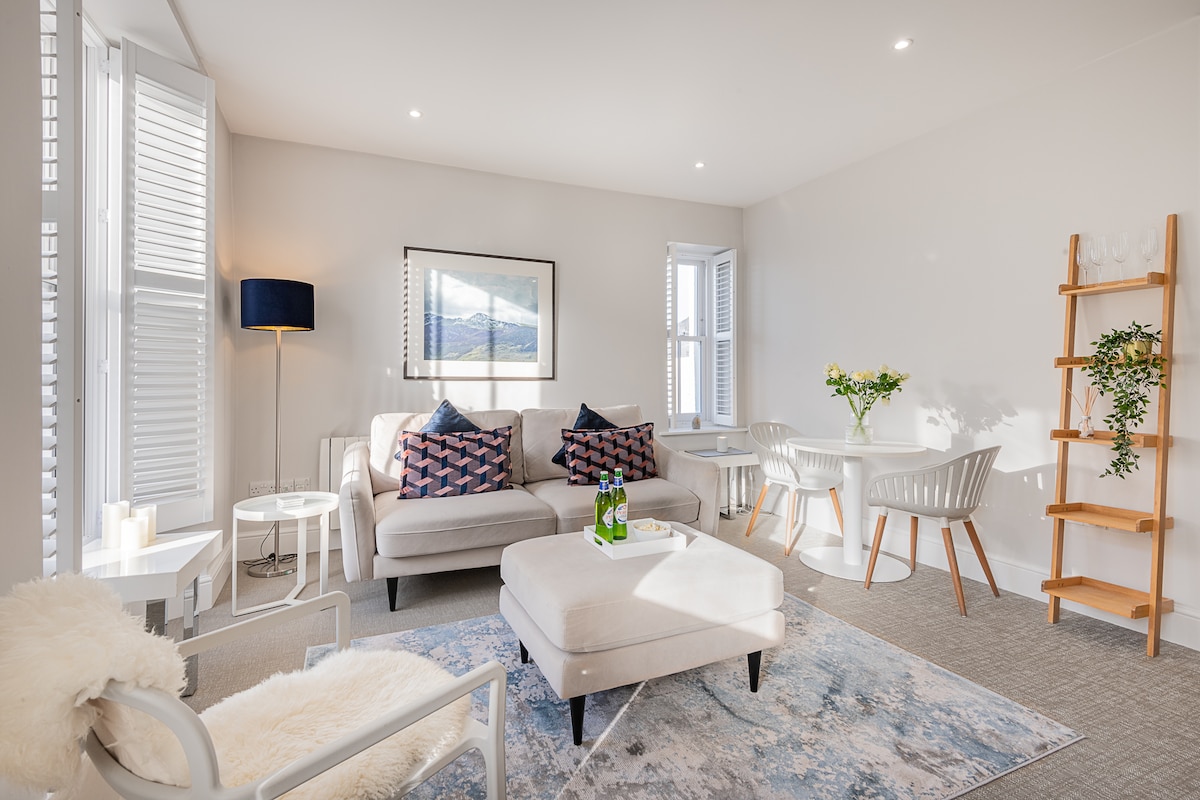 The open-plan living area features a light and airy space with two comfortable sofas adorned with decorative cushions. A round dining table for two sits beside a wooden shelving unit. Natural light fills the room through large windows, enhancing the calming atmosphere.
