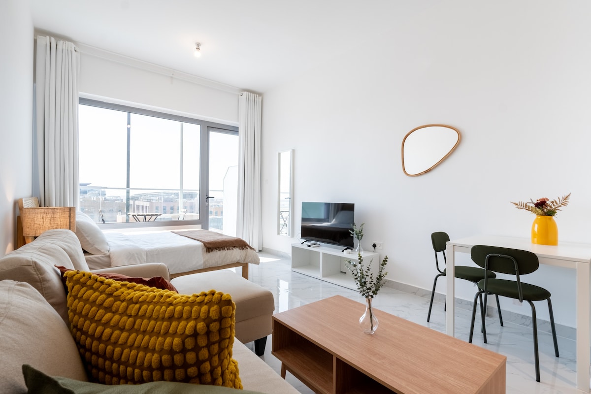 A bright living area features a comfortable sofa adorned with a textured cushion. A spacious bed is present in the background, and a dining table with chairs is positioned nearby. Large windows invite natural light, while a mirror and decorative items complete the modern decor.