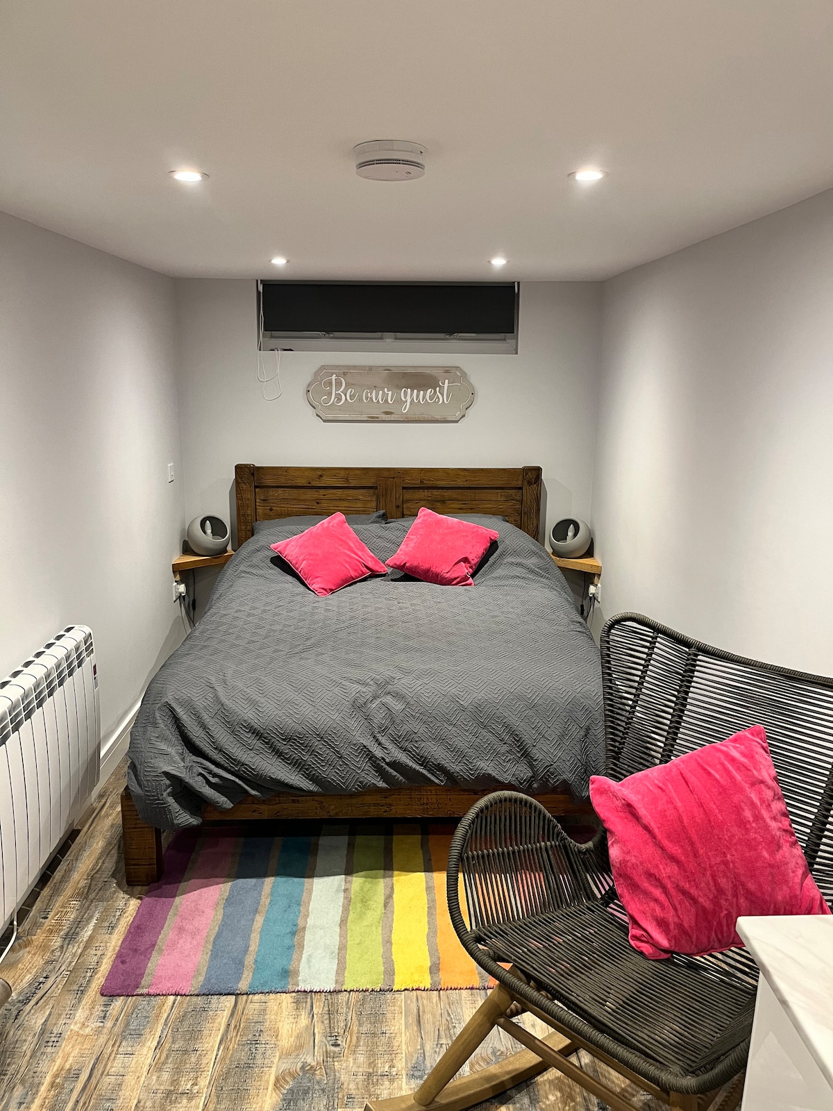 A sleeping area features a king-sized bed adorned with a dark gray cover and bright pink accent pillows. A colorful striped rug lies on the floor, complementing a wicker chair placed nearby. Soft lighting illuminates the space, contributing to a welcoming ambiance.