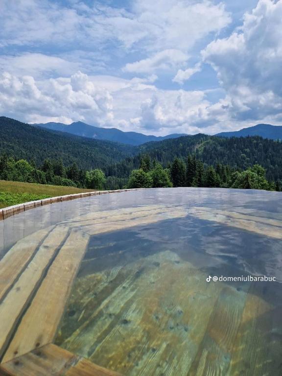 A serene outdoor pool reflects the surrounding landscape, showcasing a panoramic view of lush green hills and distant mountains. The water’s edge is lined with wooden decking, creating a harmonious blend with the natural environment under a cloudy sky.