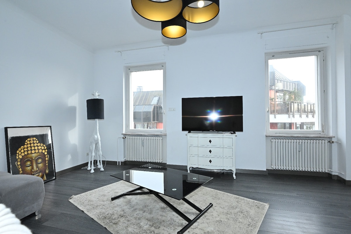 A bright living room features large windows allowing natural light, complemented by a contemporary pendant light. A sleek black and white coffee table rests on a soft area rug, while a television is positioned against a white chest of drawers. A modern floor lamp adds to the decor.