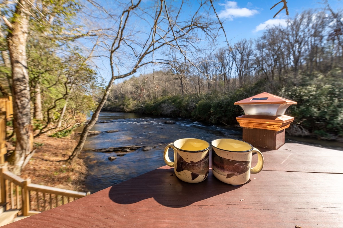 Two coffee mugs sit on a wooden table overlooking the flowing Toccoa River. Lush trees and a clear blue sky frame the serene river scene, enhancing the sense of tranquility in the surrounding nature.