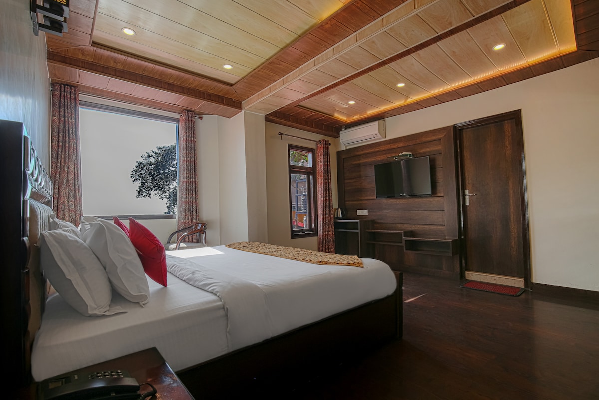 A well-appointed room features a spacious bed with neatly arranged pillows. Natural light filters through two large windows, framed by patterned curtains. The wooden ceiling and warm tones enhance the inviting ambience. A flat-screen TV is mounted on the wall opposite the bed.