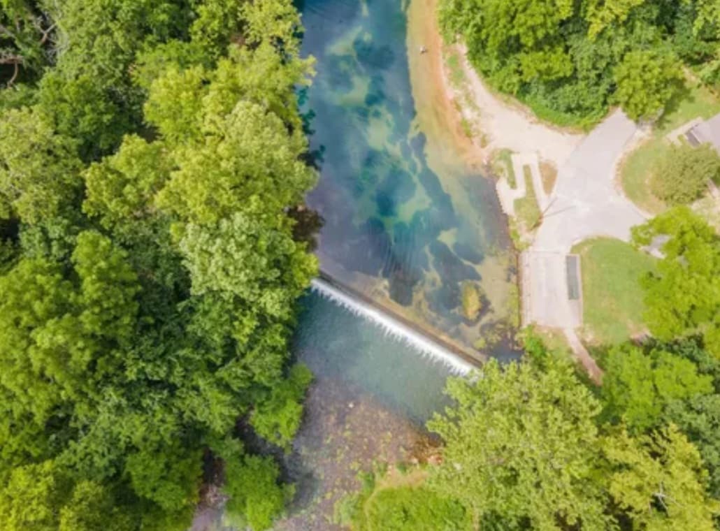 Aerial view reveals a serene waterway bordered by lush greenery. A gentle waterfall spills over a small dam, with clear water flowing beneath. Surrounding paths and picnic areas offer access to the natural beauty of the landscape.