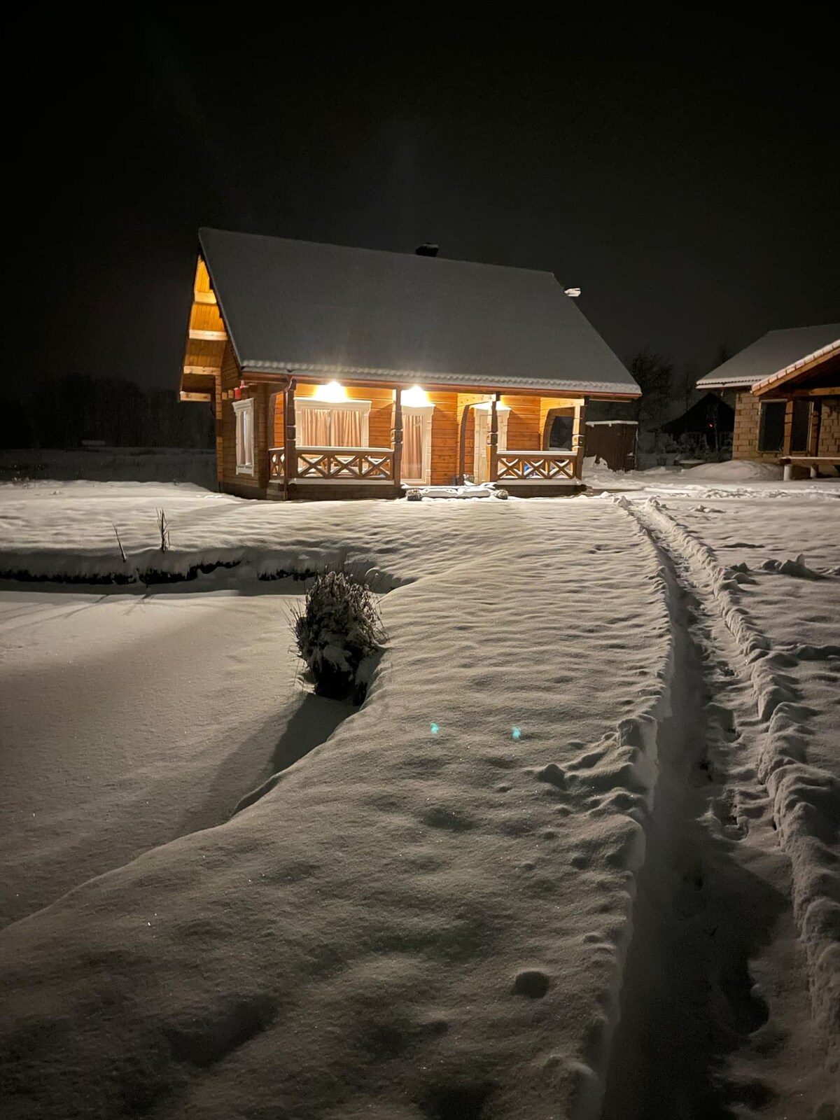 A well-lit cabin emerges from a winter landscape, surrounded by freshly fallen snow. The pathway leading to the entrance is visible, bordered by snow-covered bushes. The structure features a classic design with a sloping roof and wooden accents, conveying a serene winter ambiance.