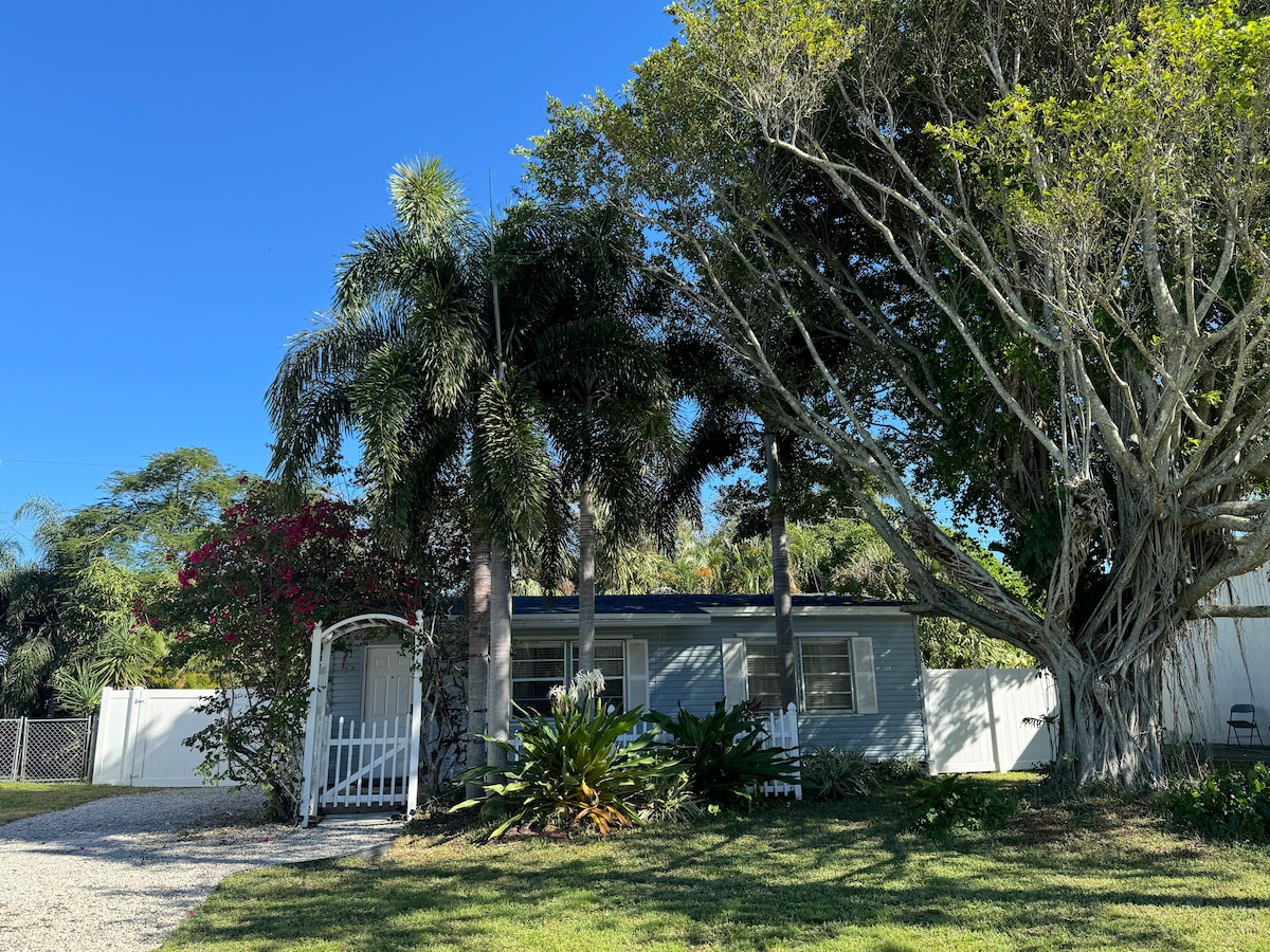 A charming exterior view of the property showcases a grey house surrounded by lush greenery. Tall palm trees and vibrant pink flowers contribute to the landscape, while a white trellis entrance adds an inviting touch. A clear blue sky creates a bright atmosphere.