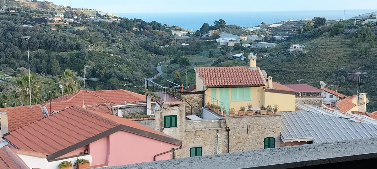 The view from the apartment showcases a scenic landscape of rooftops and rolling hills, leading toward the coastline. The clear blue sky meets the sea in the distance. The arrangement of buildings includes varied architecture, with terracotta-tiled roofs and greenery surrounding the area.