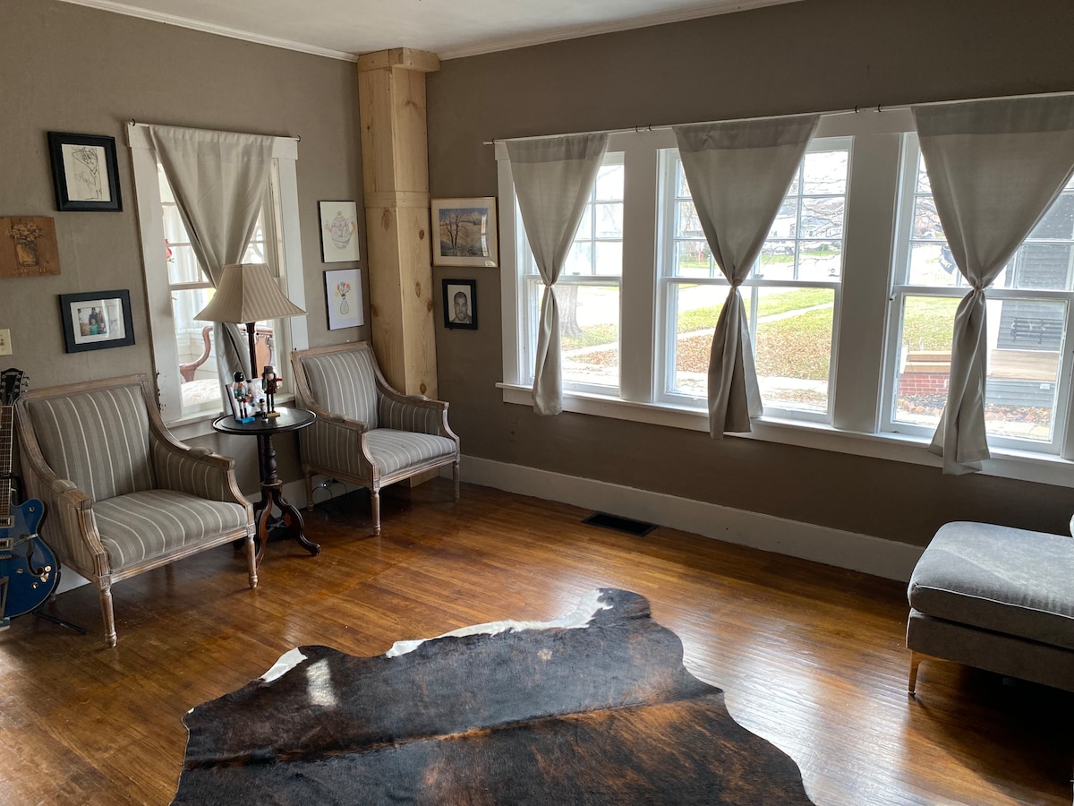 A bright living space features two striped armchairs positioned beside a small, round table, which holds a lamp and decorative items. Natural light streams through multiple windows covered by light drapes, highlighting the warm wooden floor and a decorative cowhide rug.
