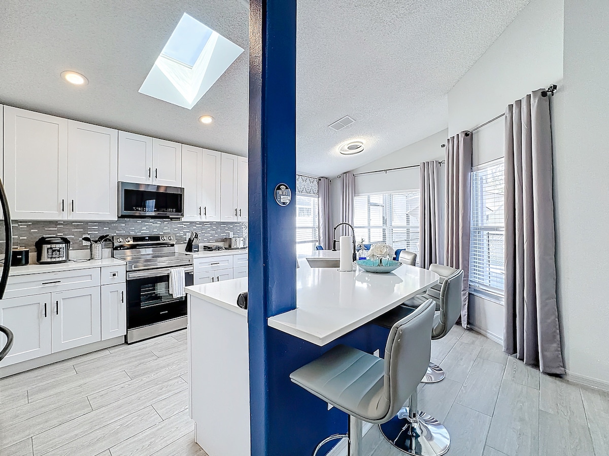 A bright, open kitchen features modern white cabinetry and stainless steel appliances. A central island with bar seating complements the space, illuminated by natural light streaming through a skylight. Large windows framed by light curtains enhance the welcoming ambiance.