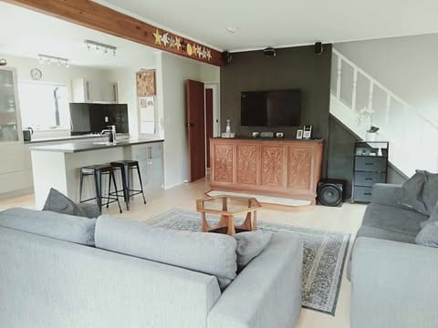 An open-plan lounge and kitchen area features a wooden bar and modern appliances. Two gray sofas sit on either side of a low wooden coffee table, while a rug anchors the seating area. Natural light enters through large windows, enhancing the spacious feel.