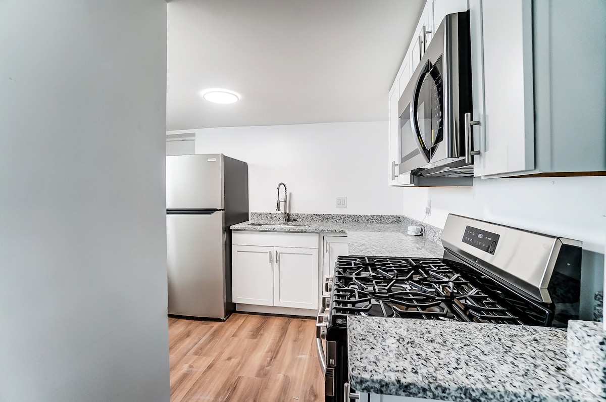 A modern kitchen is equipped with stainless steel appliances, including a refrigerator and stove. Sleek granite countertops contrast with white cabinetry. Ample natural light is provided by overhead lighting, and the flooring features a warm wood tone, enhancing the kitchen's inviting layout.