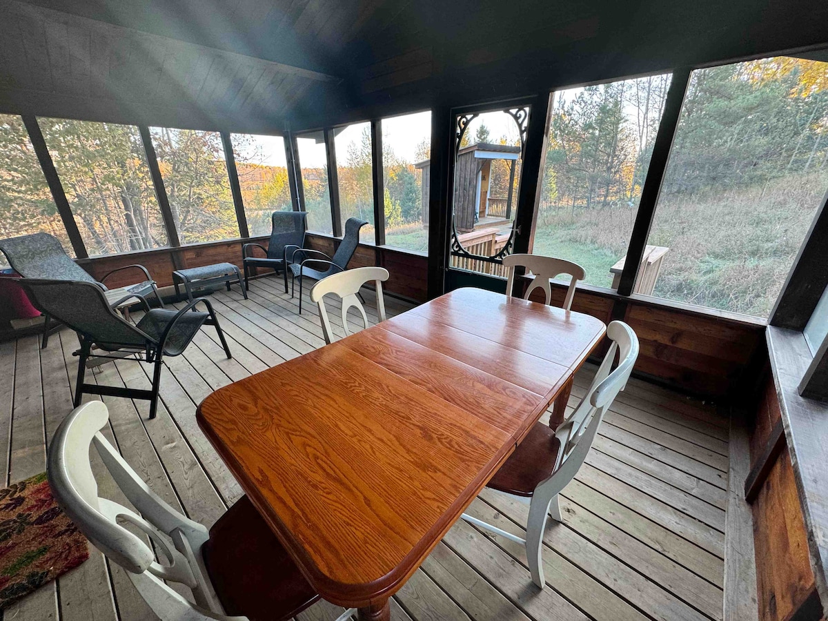 A screened porch features a wooden table surrounded by six chairs, with a blend of comfortable seating positioned for relaxation. Natural light filters through the screens, illuminating the wooden floor and offering views of the surrounding landscape.