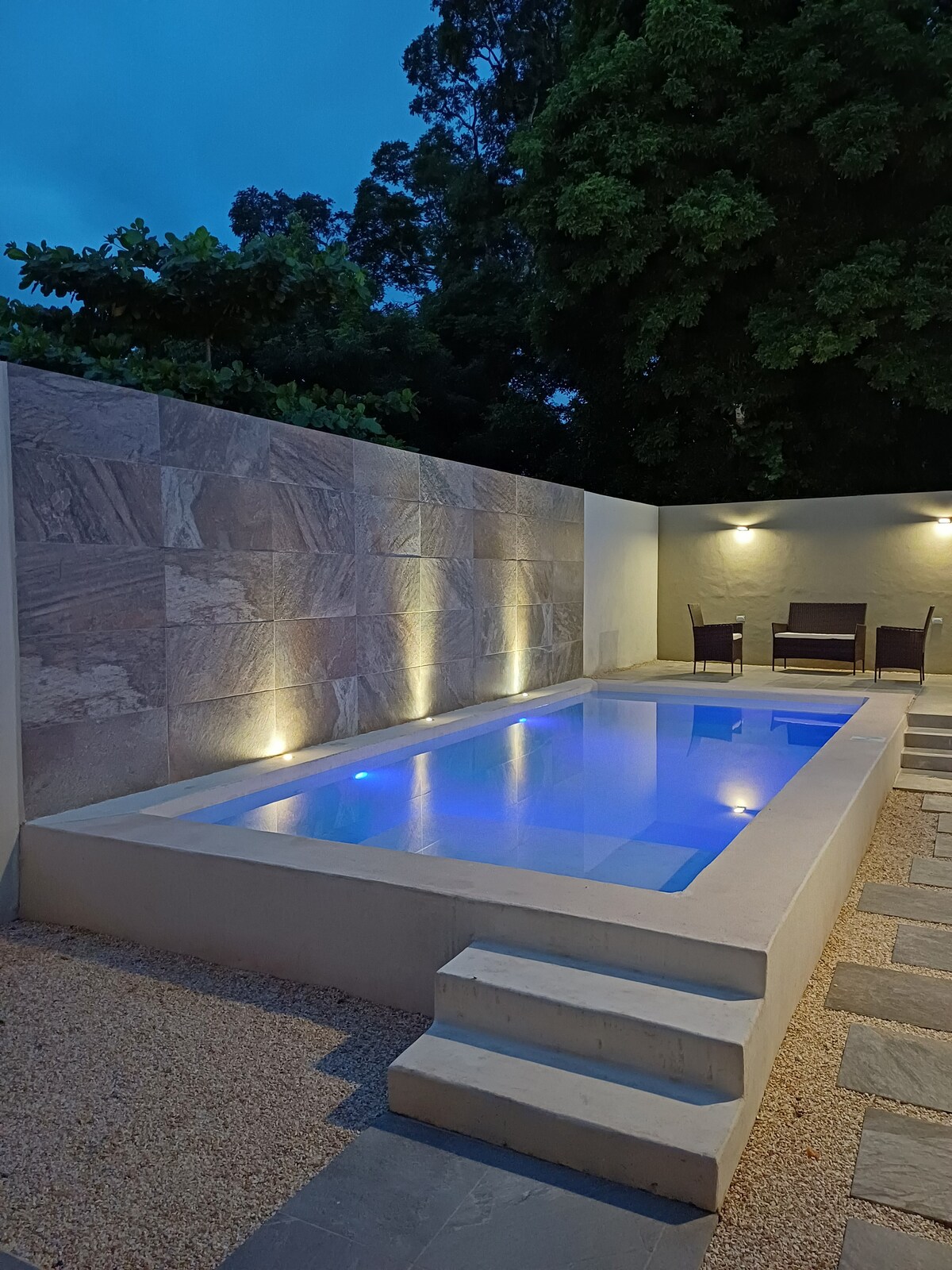 A well-lit pool area features a rectangular swimming pool with soft blue illumination, surrounded by smooth stone steps and a textured stone wall. Two outdoor chairs are set beside the pool, creating a relaxed environment in a tranquil setting.